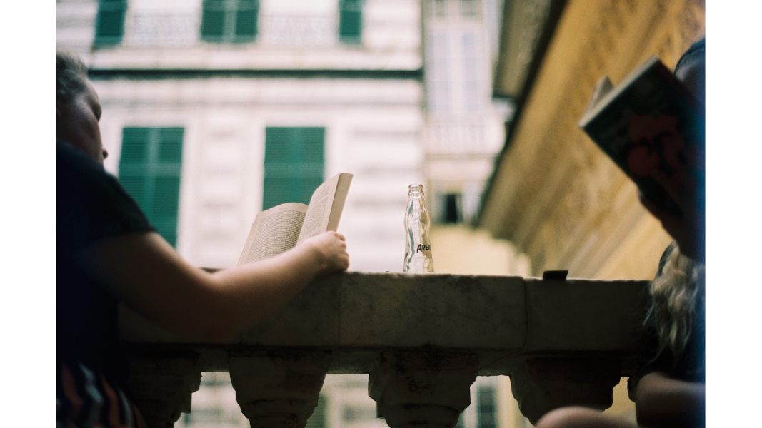 a film photo looking up from a low angle towards 2 friends reading on a balcony, an empty bottle of aperal spritz between them.