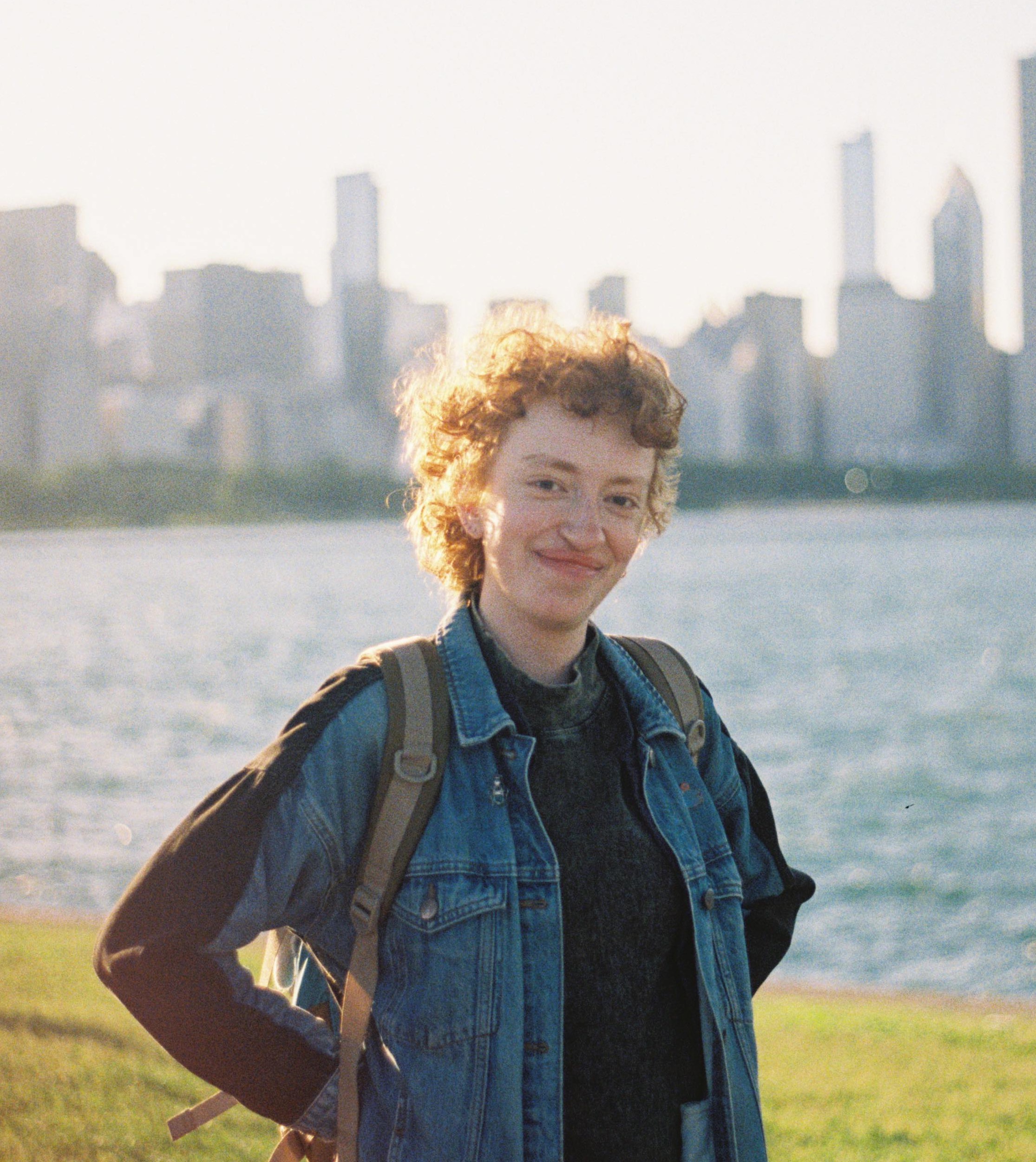 a photograph of Storm, a white person with short ginger hair, holding a camera in front of some rolling hills.